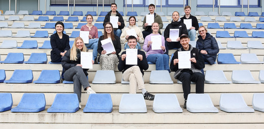 mehrere Personen sitzen auf Besucher*innenrängen eines Stadions und halten Urkunden hoch
